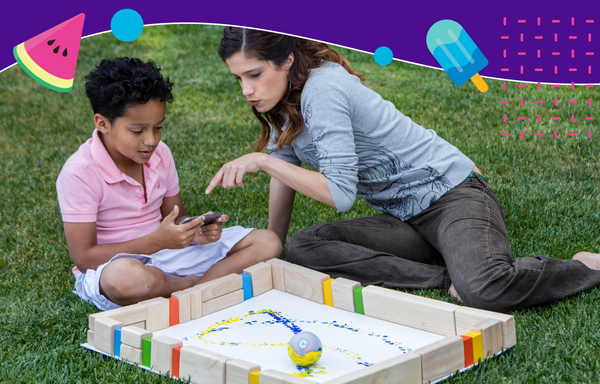A boy and woman sit in the grass outside painting with a Sphero robot. 