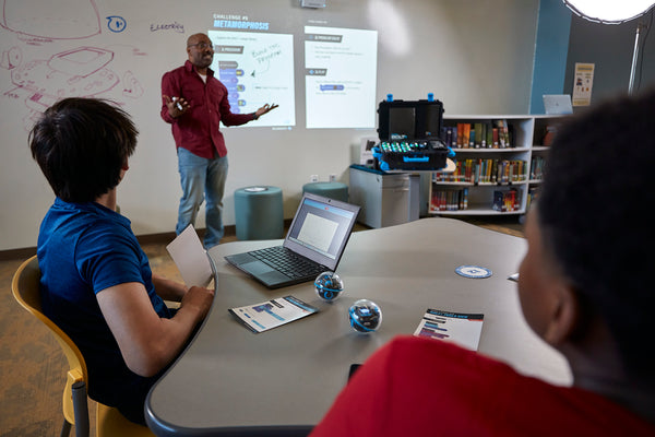 A teachers stands in front of his class with a projector walking his students through a lesson with Sphero BOLT+. The students are looking on with their Chromebooks at desks with their robots.