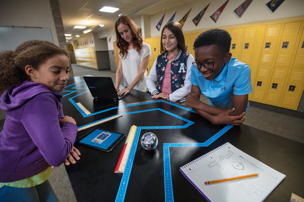 Three students and their teacher stand around a table with a maze made from tape as they navigate their Sphero BOLT from one end to the other. 