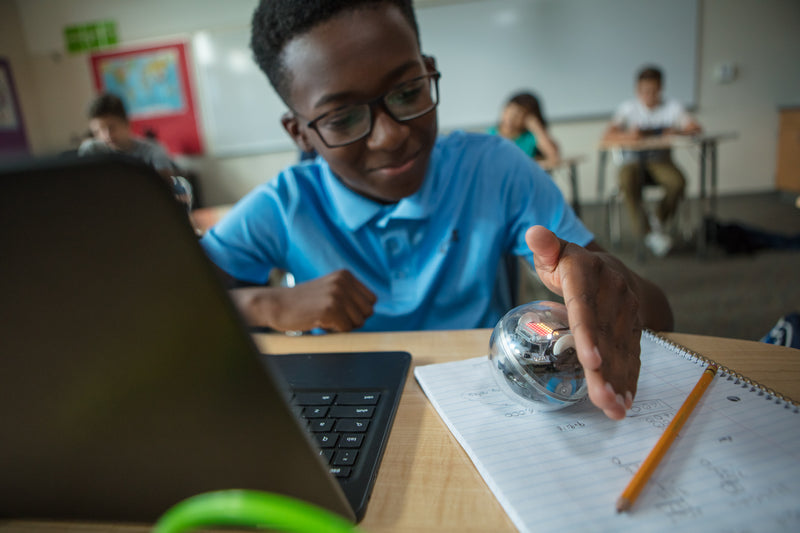 A boy sits at his desk in a classroom working on a computer with Sphero BOLT.