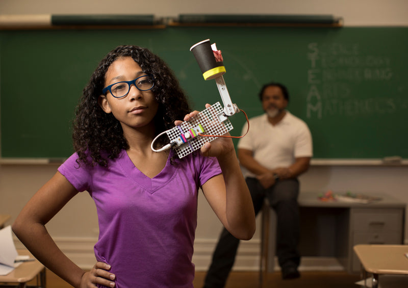 A teenage girl shows off her littleBits invention in her classroom while her teacher look on from the background.