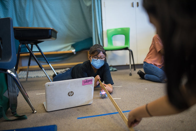 A girl in a mask lies on the floor of her classroom measuring distanced with a yard stick while programming her Sphero BOLT on a laptop.