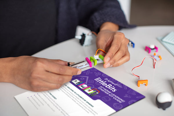 A person snaps together pink and green littleBits at a white table with a getting started guide nearyby.