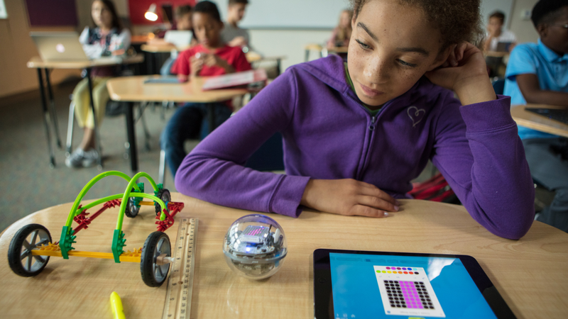 A girl in a purple sweatshirt sits at her desk coding her Sphero BOLT on a tablet.