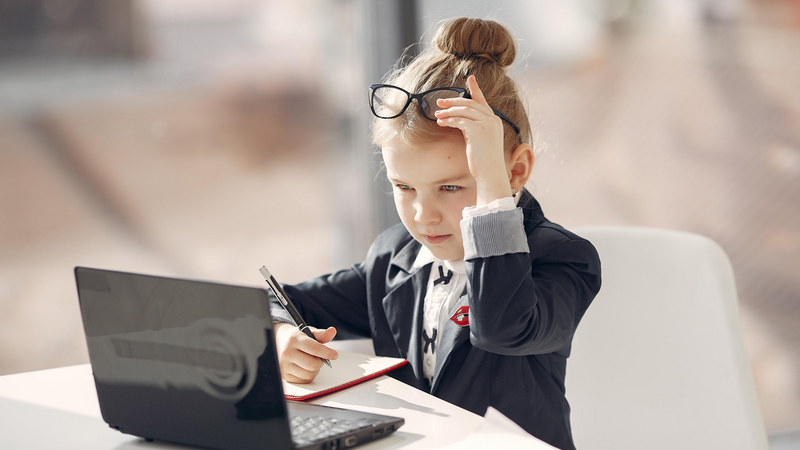 A young girl with eyeglass on top of her head sits at a table in front of her laptop.