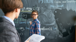 A girl in glasses writes code on a chalkboard while her classmate reads something to her from notebook.