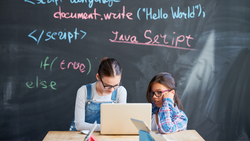 Two girls work together on a laptop; Javascript text is on the chalkboard behind them.