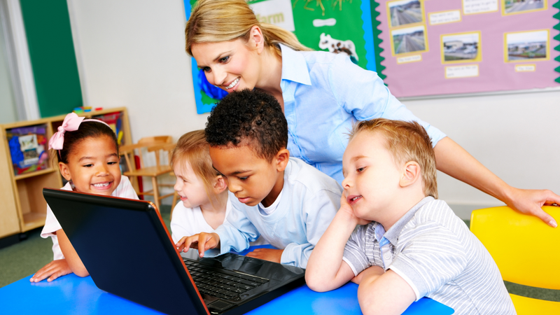 A teacher works with four younger students on a laptop in a colorful classroom. Cybersecurity education is important for teachers and students to learn.