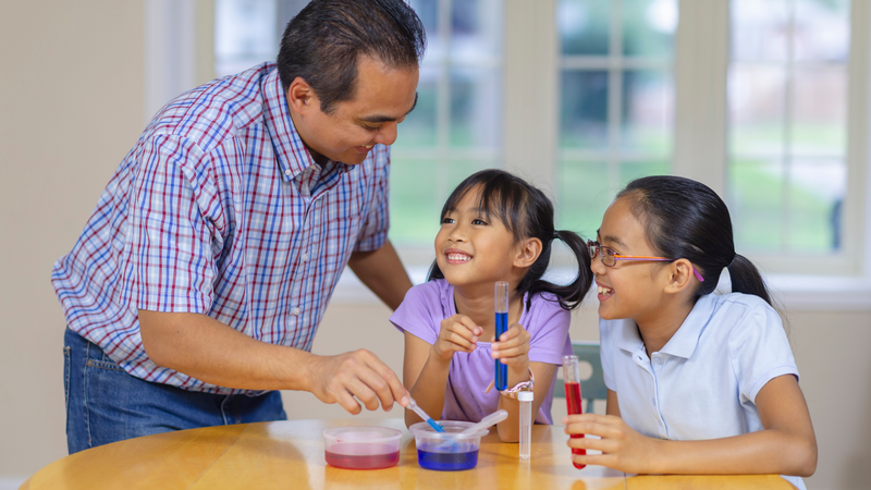 A father and his two daughters work on a science activity at the kitchen table.