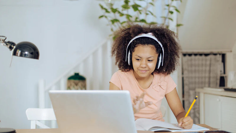 A girl does her school work on a laptop while wearing headphones at home. This is an example of a hybrid-learning environment.