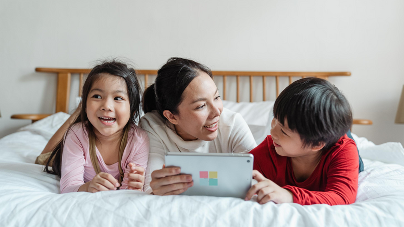 A mom and her daughter and son lay on a bed looking at a tablet together.