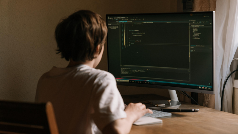 A boy sits at a computer at a desk working on a program.
