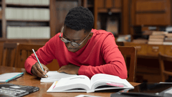 A male student takes written notes while reading from a textbook at a desk.