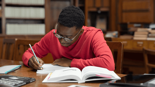 A male student takes written notes while reading from a textbook at a desk.