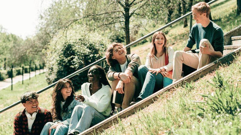 A group of teenagers sits on stairs chatting and laughing with each other. STEM Education is just one foundational way for kids and teens to learn importance social interactions in school.