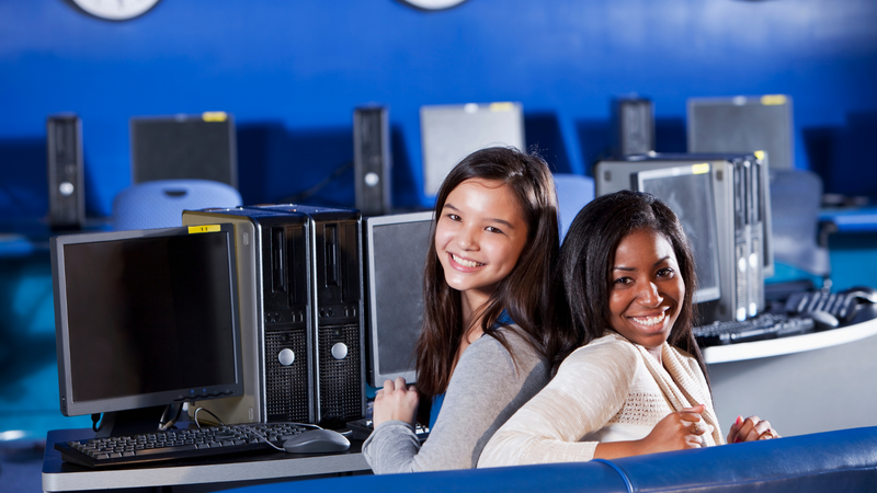 Two teenager girls sit together back to back in a computer lab at school.