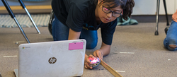 A student kneels down next to a computer with a ruler and the  Sphero BOLT Coding Robot in hand.