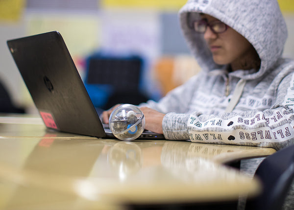 A kid with a white hoodie on a laptop and sitting next to a toy robot ball. 