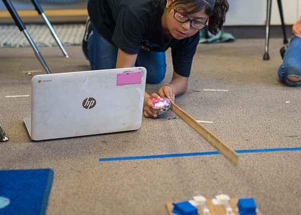 Girl with laptop kneeling on the floor with STEAM robot and ruler.
