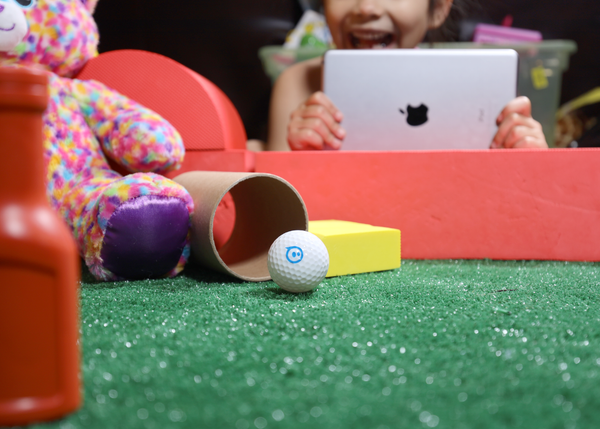 A hand driving a toy robot soccer ball using block-based coding through the Sphero Edu app.