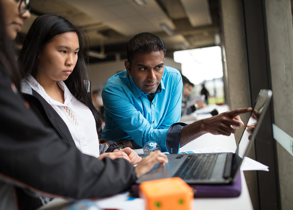 Teacher pointing to high school student's laptop screen beside a STEM robot.